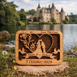 May include: Wooden wedding announcement with a heart-shaped cutout featuring kissing figures. The announcement reads "Save The Date" at the top, with the names "Kate & Michael" and the date "5 October, 2029" below. The background shows a castle and lake.