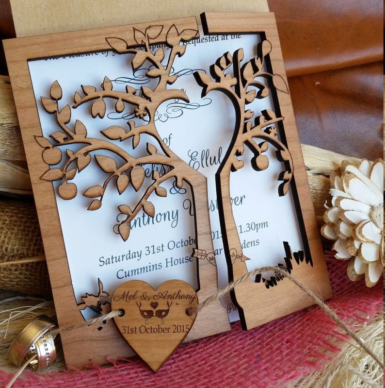 May include: A laser-cut wooden wedding invitation with a tree design. The invitation features a heart-shaped tag with the names "Mel & Anthony" and the date "31st October 2015". The invitation is made of light brown wood and includes the wedding details.