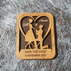 May include: A wooden save the date card with a heart cutout featuring a couple silhouette. The card is engraved with the names "Jill & Steven" and the date "6 September 2015".