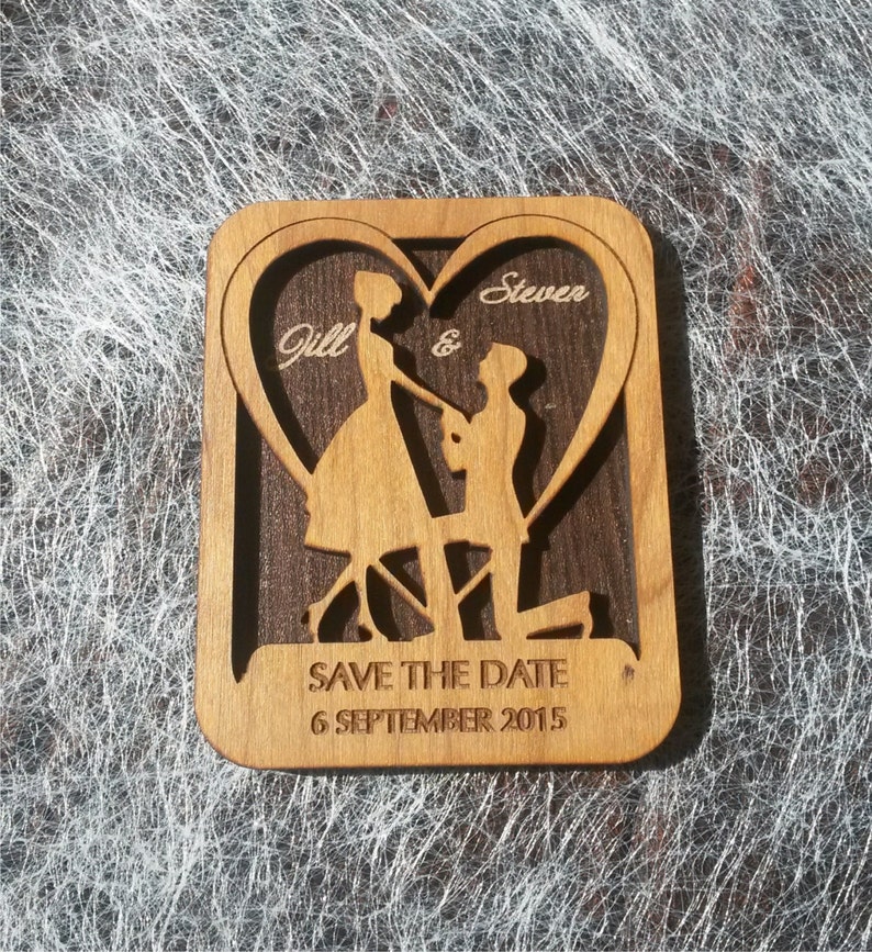May include: A wooden save the date card with a heart cutout featuring a couple silhouette. The card is engraved with the names "Jill & Steven" and the date "6 September 2015".