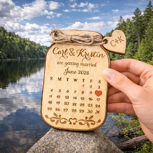 May include: Wooden wedding calendar in the shape of a jar, with the names "Carl & Kristin" and the text "are getting married June 2028." A small wooden heart with the initials "C+K" hangs from the top. The calendar has a heart marking the 16th of June.