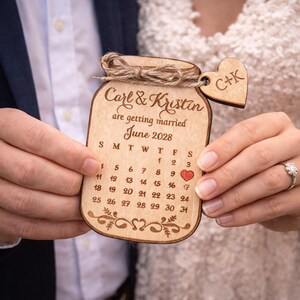 May include: A wooden wedding announcement shaped like a jar, with the names "Carl & Kristin" and the date "June 2028" engraved. A small heart marks the wedding date on the calendar. A small wooden heart with the initials "C+K" is attached.