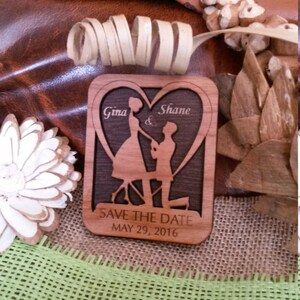 May include: A wooden save the date card with a heart-shaped cutout featuring a couple in silhouette. The card is engraved with the names "Gina & Shane" and the date "MAY 29, 2016".