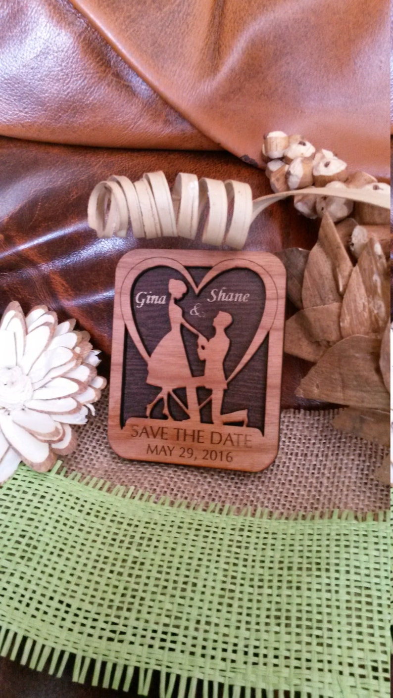 May include: A wooden save the date card with a heart-shaped cutout featuring a couple in silhouette. The card is engraved with the names "Gina & Shane" and the date "MAY 29, 2016".