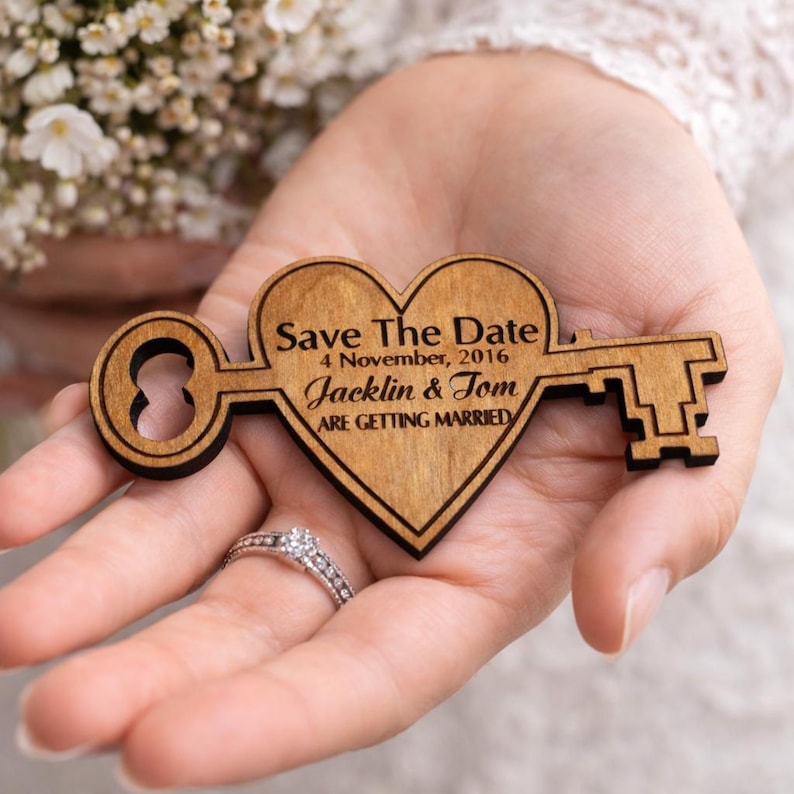 May include: A wooden heart-shaped key with the text "Save The Date" and the names "Jacklin & Tom" is held in a hand. The key is a light brown color with a darker brown outline. The text "ARE GETTING MARRIED" is also visible.