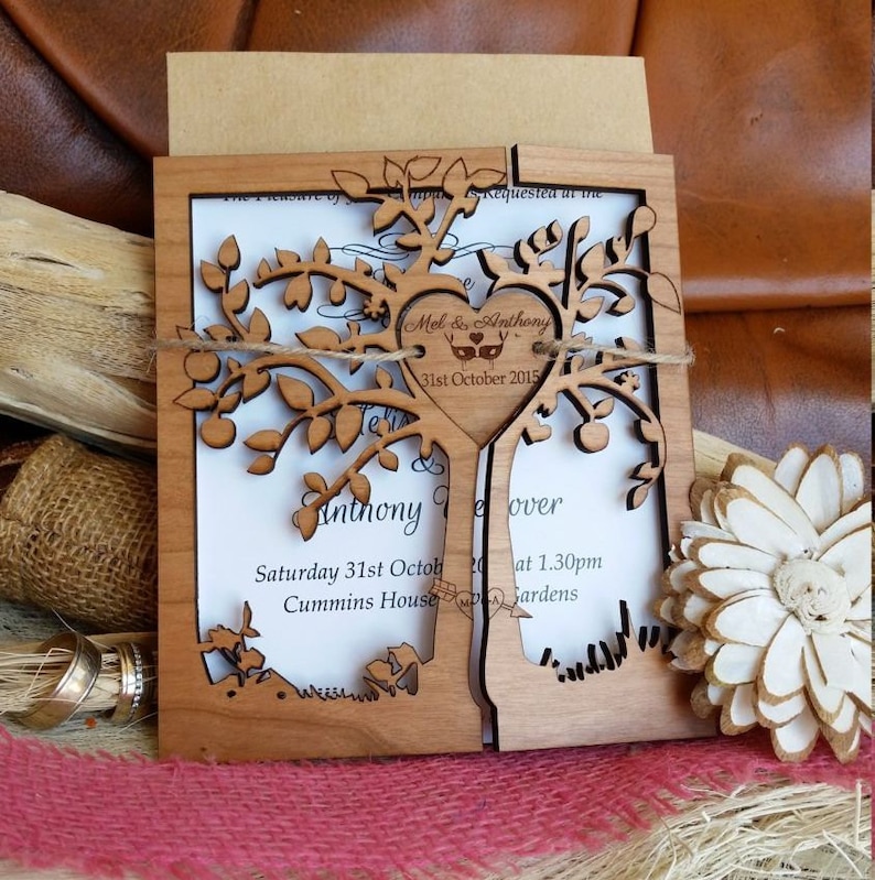 May include: A laser-cut wooden wedding invitation featuring a tree design. The invitation includes the names "Mel & Anthony" and the date "31st October 2015". The invitation is tied with twine and has a kraft paper envelope.
