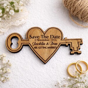 May include: Wooden heart-shaped key with the text "Save The Date" and wedding details. The key is light brown with a dark brown outline. Gold wedding rings and white flowers are also in the image.