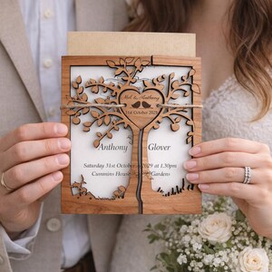 May include: A wooden wedding invitation with a tree design. The invitation features the names "Mel & Anthony" and the date "31st October 2029". The invitation is held by two people, with a bouquet of white roses in the background.