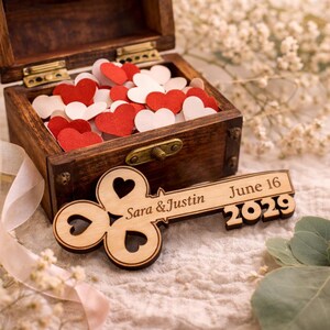 May include: A wooden key-shaped wedding favor with the names "Sara & Justin" and the date "June 16 2029" engraved on it. A small wooden box filled with red and white heart-shaped confetti is in the background.