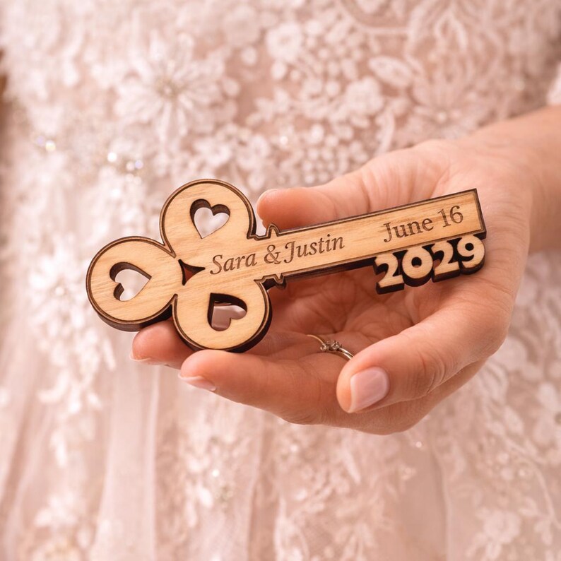 May include: A wooden key-shaped wedding favor with the names "Sara & Justin" and the date "June 16 2029" engraved on it. The key has heart-shaped cutouts and is held in a person's hand.