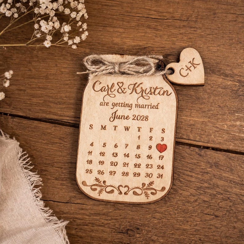 May include: A wooden wedding calendar in the shape of a jar, with the names "Carl & Kristin" and the date "June 2028" engraved. A small wooden heart with "C+K" hangs from the top. The calendar has a heart on the 17th.