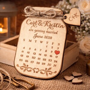 May include: Wooden wedding calendar in the shape of a jar, engraved with "Carl & Kristin are getting married June 2028." A small wooden heart with "C+K" hangs from the top. The calendar has a red heart marking June 9th.
