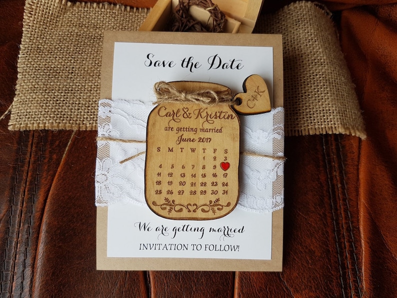 May include: A rustic wedding invitation featuring a wooden jar-shaped calendar with the names "Carl & Kristin" and the date "June 2017". The invitation includes lace, twine, and a wooden heart with initials.