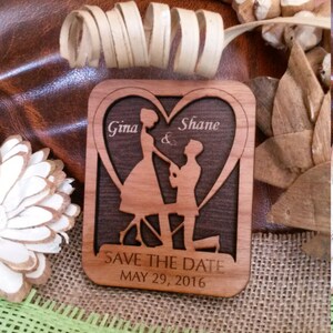 May include: Wooden save the date card with a silhouette of a couple in a heart shape. The card is engraved with the names "Gina & Shane" and the date "MAY 29, 2016".