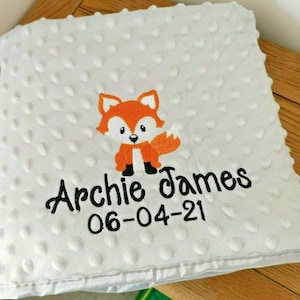 May include: A white baby blanket with a small orange fox embroidered on it. The blanket has a textured surface and the words "Archie James 06-04-21" are embroidered below the fox.