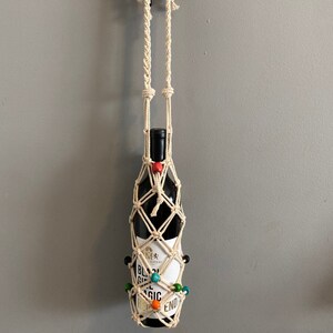 Op de afbeelding: Een macram&eacute; wijnfleshouder met een wit koord en kleurrijke kralen. De fleshouder hangt aan een haak aan een muur. De fles is gelabeld "Black Girl Magic" en bevat rode wijn.