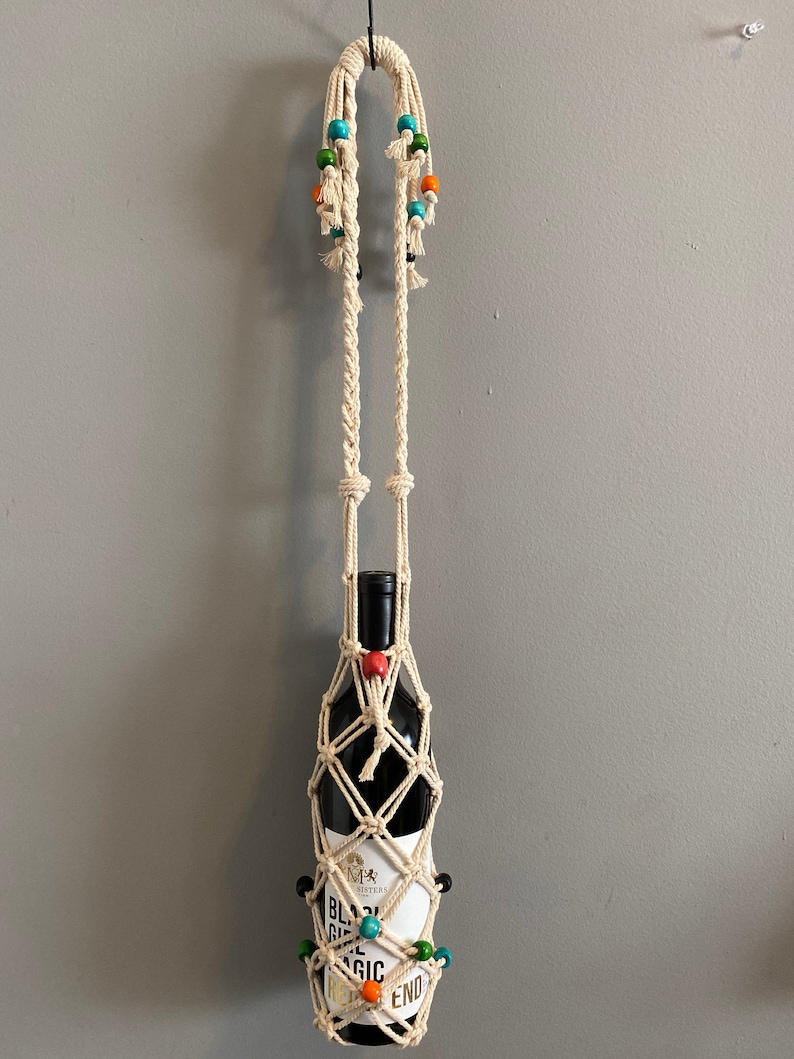 Op de afbeelding: Een macram&eacute; wijnfleshouder met een wit koord en kleurrijke kralen. De fleshouder hangt aan een haak aan een muur. De fles is gelabeld "Black Girl Magic" en bevat rode wijn.