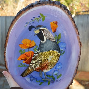 May include: A hand-painted wooden slice with a purple background features a colorful quail with a crown of blue flowers. The quail is surrounded by orange and yellow flowers and green leaves. The artist's signature is in the bottom right corner.