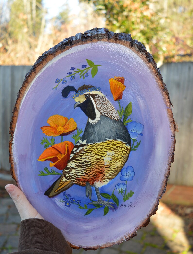 May include: A hand-painted wooden slice with a purple background features a colorful quail with a crown of blue flowers. The quail is surrounded by orange and yellow flowers and green leaves. The artist's signature is in the bottom right corner.