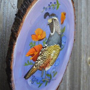 May include: A hand-painted wooden slice with a purple background features a colorful California Quail with a crown of blue flowers. The bird is surrounded by orange poppies and blue forget-me-nots.