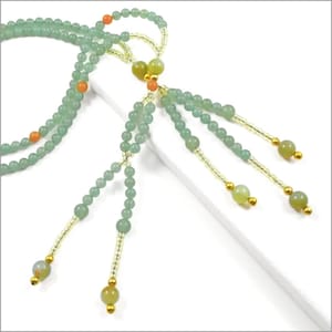 May include: A delicate necklace featuring light green beads and accents of orange and gold. The necklace has multiple strands and beaded tassels, creating an elegant and intricate design. The beads are small and round, with a subtle sheen.