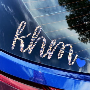 May include: A blue car with a rear window sticker that says "Rhm" in a leopard print font with a blue heart next to it.