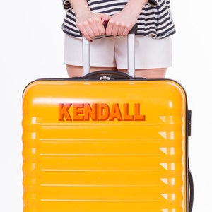 May include: A bright yellow suitcase with black wheels and a black handle. The suitcase has the word "KENDALL" printed in red and orange letters on the front.