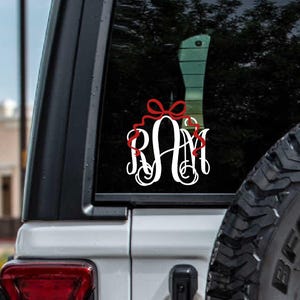 May include: White monogram decal with the letters "RAM" in a decorative script, adorned with a red bow. The decal is affixed to the rear window of a white vehicle, with a spare tire visible on the right.