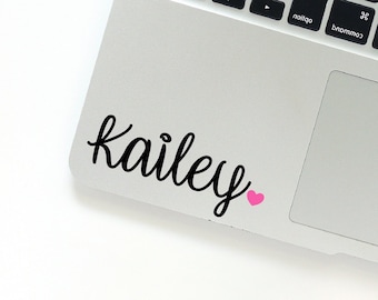 Pode incluir: Um laptop prateado com um teclado preto e um touchpad preto. O laptop tem um adesivo preto e branco com o nome "Kailey" e um coração rosa.