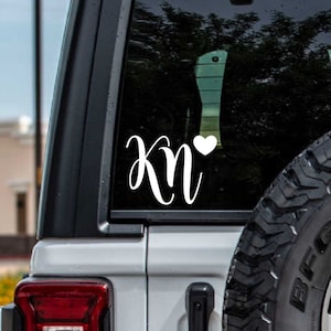 May include: White vinyl decal with the letters "KN" in a cursive font and a small white heart to the right of the letters. The decal is on the back window of a gray Jeep.