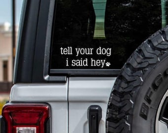 May include: White car with a black tyre and a sticker on the rear window that says "tell your dog i said hey" with a paw print.