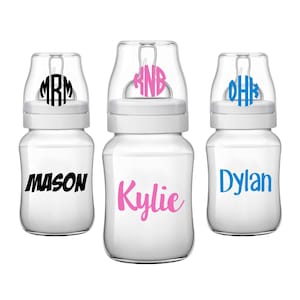 May include: Three clear baby bottles with personalized labels. The bottles have white caps with a monogram on each. The labels have the names "Mason", "Kylie", and "Dylan" written in black, pink, and blue lettering.