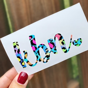 May include: White sticker with the word "kbn" in a colorful leopard print design. The letters are outlined in black and filled with neon pink, blue, yellow, and green spots. The sticker is held by a hand with red nail polish.