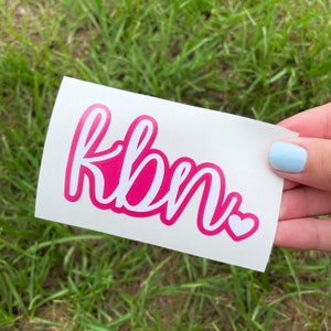 May include: A white sticker with the word "kbn" written in pink cursive lettering with a white outline. A small white heart is to the right of the word.