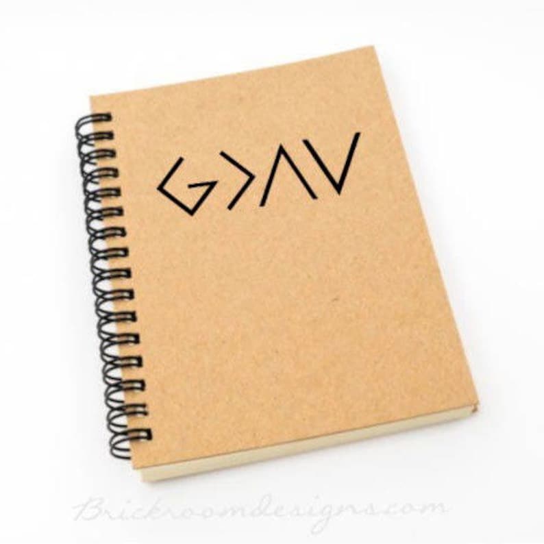 May include: A brown spiral bound notebook with a black graphic of the words "God is greater than" written in a stylized font.