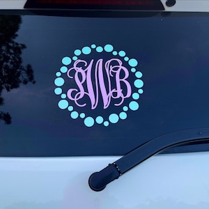 May include: A custom car decal featuring a monogram in lavender, surrounded by a circle of light blue dots. The decal is applied to a dark surface, with a car wiper visible at the bottom.