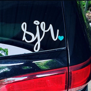 May include: A white vinyl decal with the word "sju" in cursive lettering with a small turquoise heart next to it. The decal is on the back window of a black car.