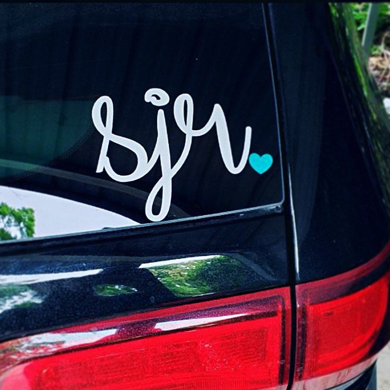 May include: A white vinyl decal with the word "sju" in cursive lettering with a small turquoise heart next to it. The decal is on the back window of a black car.