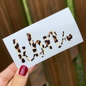 May include: A white sticker with the word "KBN" in a cursive font. The letters are filled with a leopard print pattern in shades of brown and tan. A small heart with the same pattern is next to the last letter.