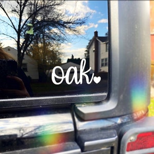 Pode incluir: Adesivo de vinil branco com a palavra "oak" em letra cursiva e um pequeno cora&ccedil;&atilde;o ao lado. O adesivo est&aacute; na janela traseira de um ve&iacute;culo.