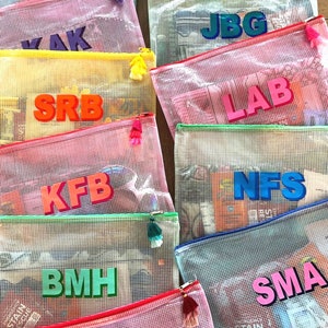 May include: A collection of colorful mesh zippered pouches with different names written on them in bold letters. The pouches are made of a clear mesh material and have a bright colored zipper and tassel.