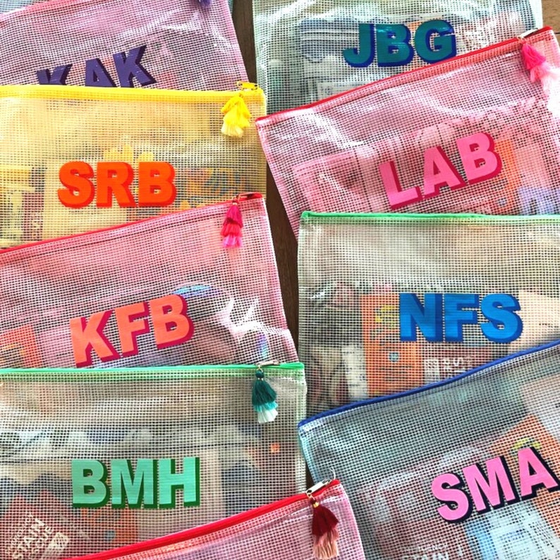 May include: A collection of colorful mesh zippered pouches with different names written on them in bold letters. The pouches are made of a clear mesh material and have a bright colored zipper and tassel.