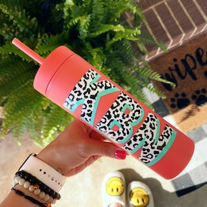 May include: A pink tumbler with a black and white leopard print design and the letters "KB" in turquoise. The tumbler has a straw.