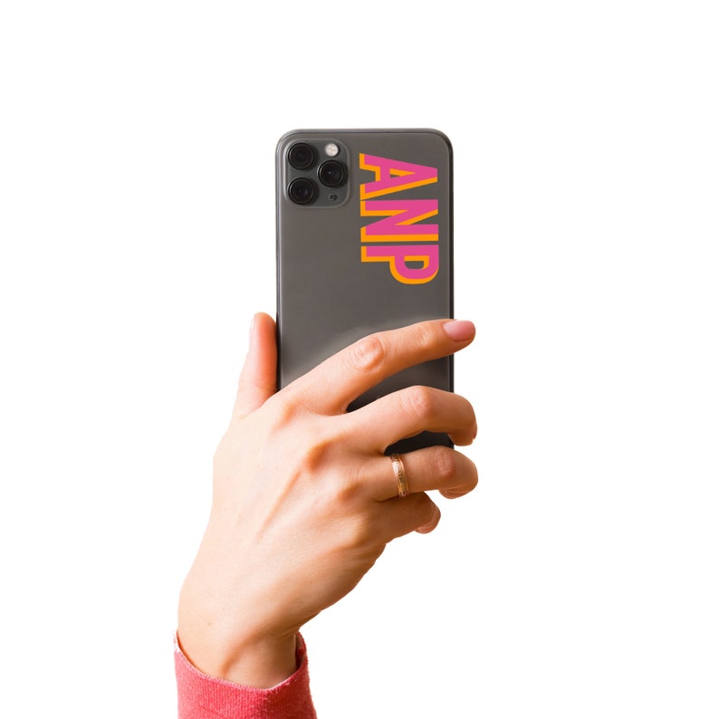 Puede incluir: Una mano sosteniendo un tel&eacute;fono inteligente negro con una pegatina rosa y naranja que dice "ANP" en la parte posterior.