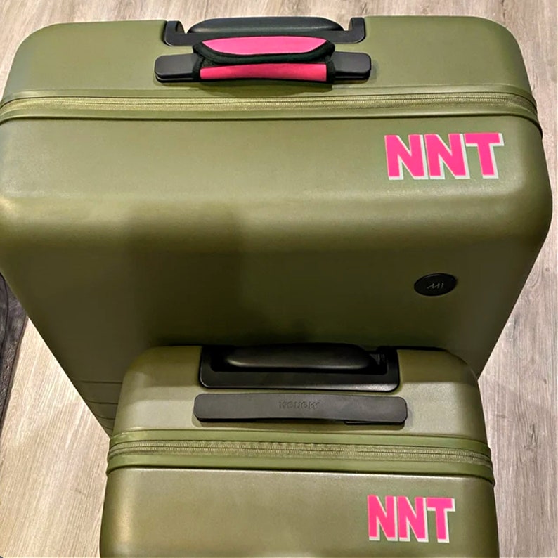 May include: Two olive green suitcases with black handles and pink lettering that says "NNT" on each suitcase.