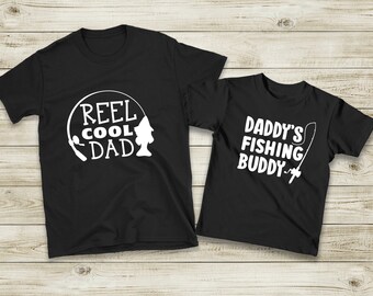 Könnte beinhalten: Zwei schwarze T-Shirts mit weißem Text und Grafiken. Das größere Shirt sagt "Reel Cool Dad" mit einer Angelrute und einem Fisch-Motiv. Das kleinere Shirt sagt "Daddy's Fishing Buddy" mit einer Angelrute.
