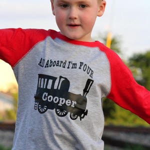 Puede incluir: Una camiseta de manga larga gris y roja con un diseño de tren negro y el texto "All Aboard I'm Four" y "Cooper".