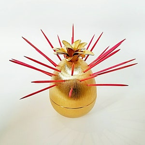 May include: A gold pineapple-shaped toothpick holder with red toothpicks sticking out of it.