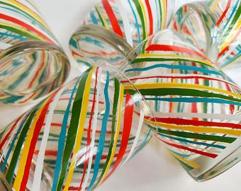 May include: Set of six clear glass tumblers with a colorful striped design. The stripes are red, yellow, green, blue, and white. The stripes are diagonal and create a swirling pattern.