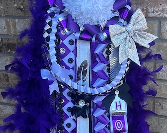 Puede incluir: Mum de regreso a casa de color morado y blanco con un lazo plateado, una cinta a cuadros blanco y negro y una pluma blanca. La mum tiene un letrero que dice "Junior" y un letrero que dice "Homecoming".